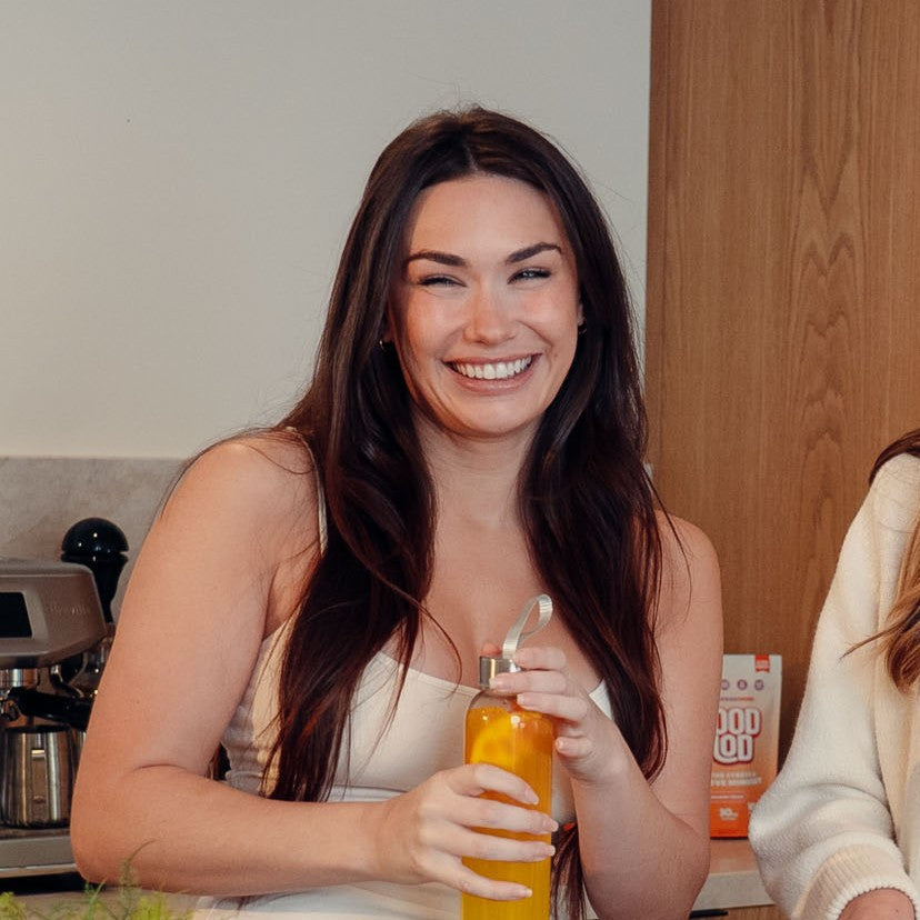 Smiling woman with long dark hair holding a bottle of orange Mood Mod saffron drink in a kitchen, with a Mood Mod pouch in the background