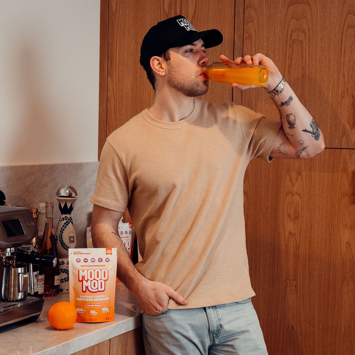 Man in a Mood Mod cap drinking an orange saffron drink from a bottle, with a Mood Mod Orange Cream pouch and fresh orange on the counter