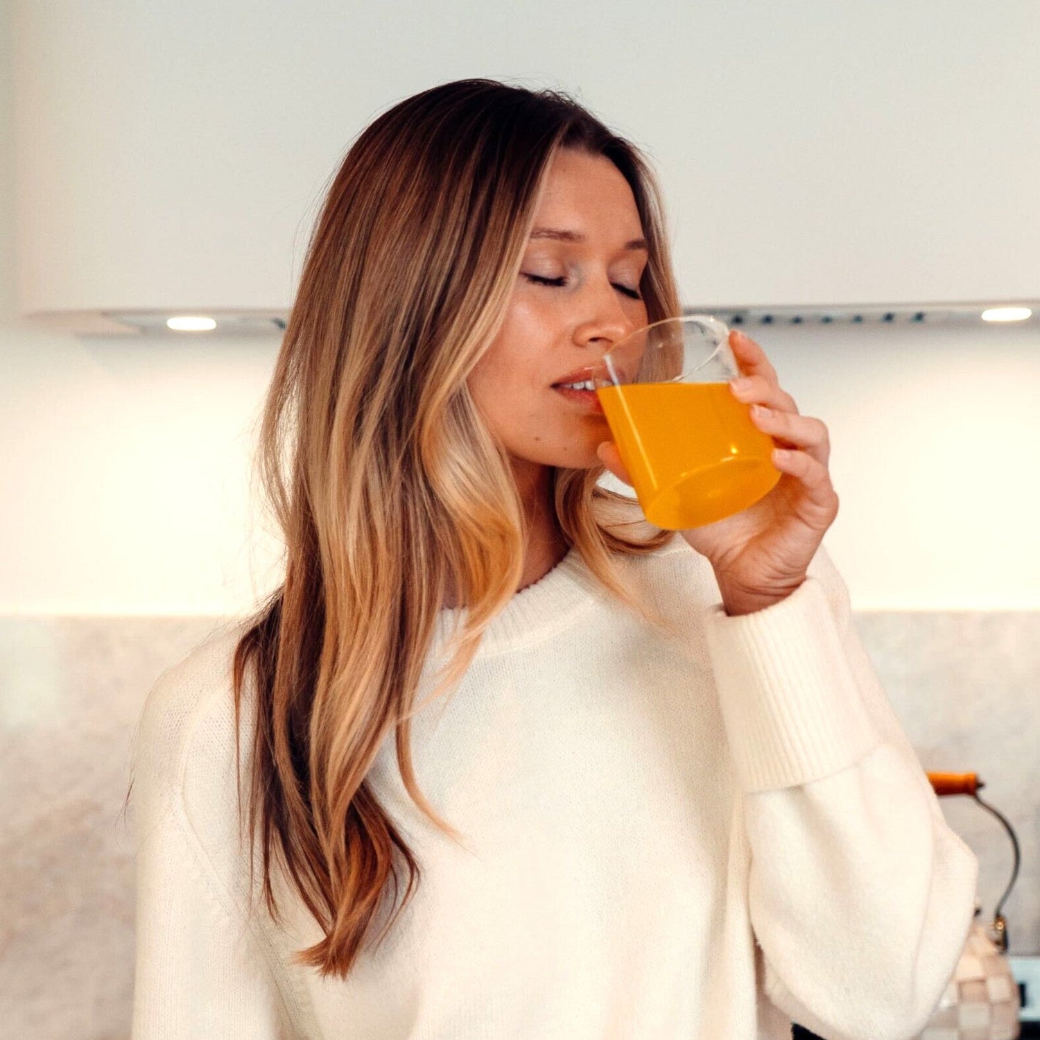Woman with long blonde hair in a cream sweater drinking an orange Mood Mod saffron drink from a glass in a bright kitchen