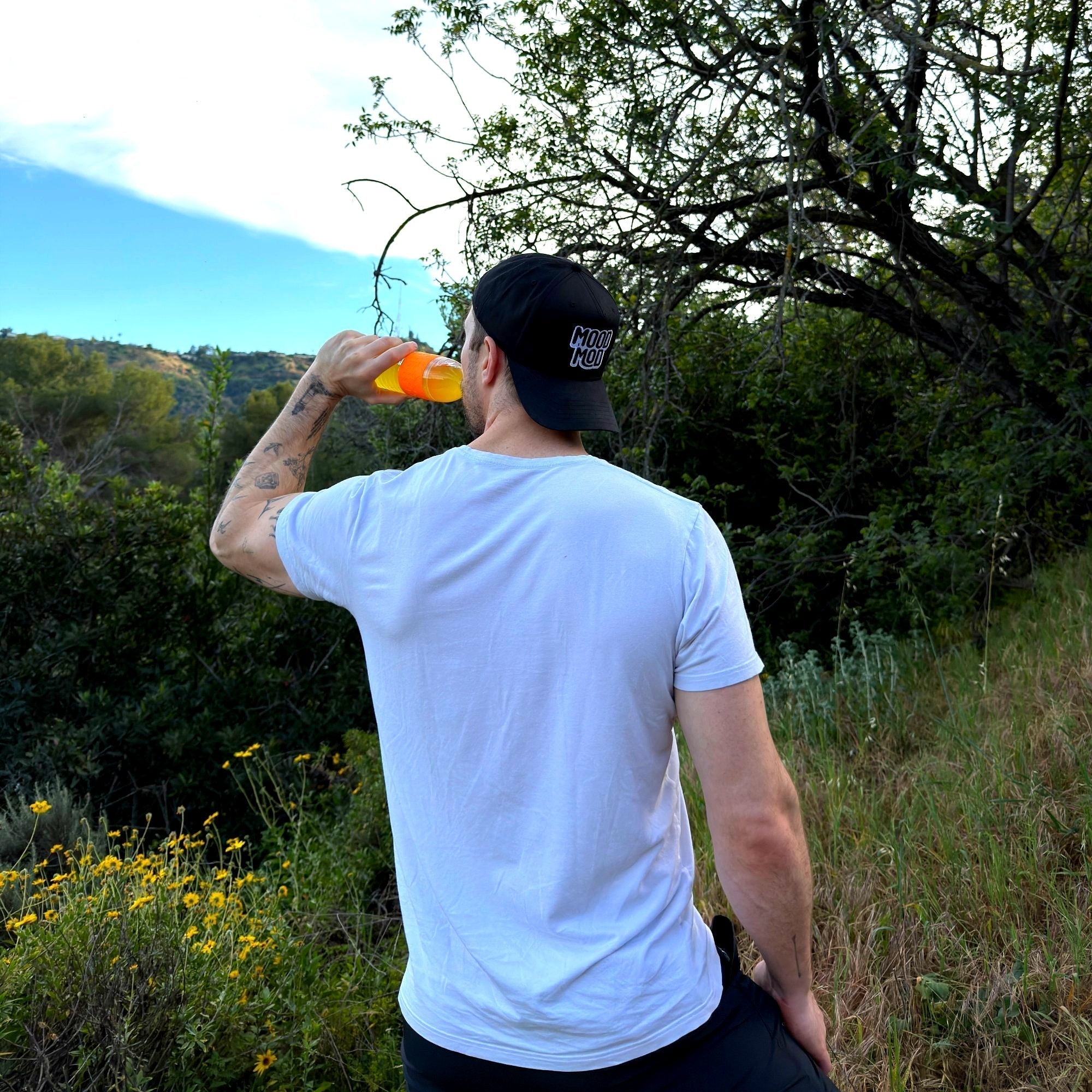 Man in a white t-shirt and black cap drinking from a bottle while overlooking a green valley