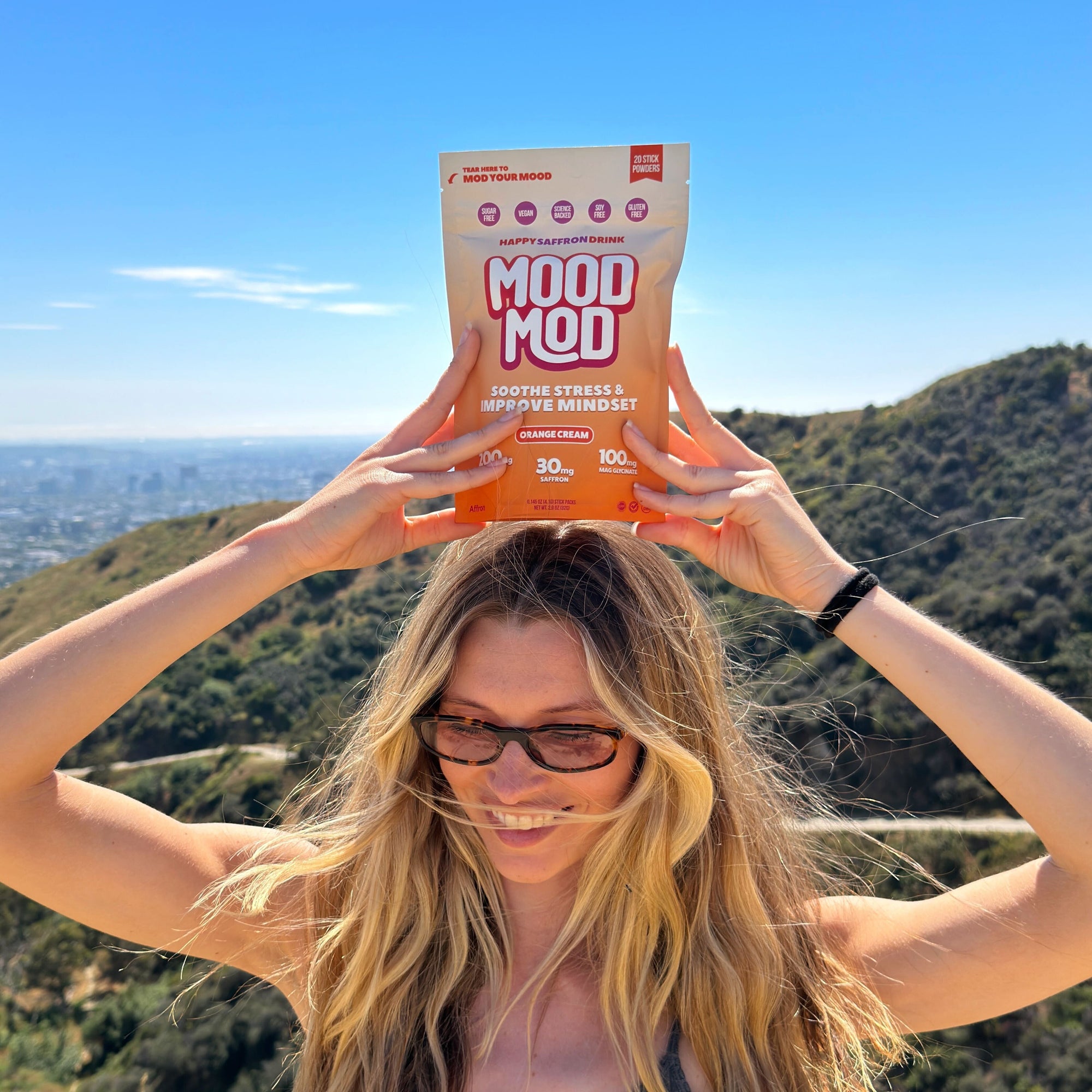 Smiling woman holding Mood Mod Happy Saffron Drink pouch above her head on a hilltop trail with LA cityscape in the background