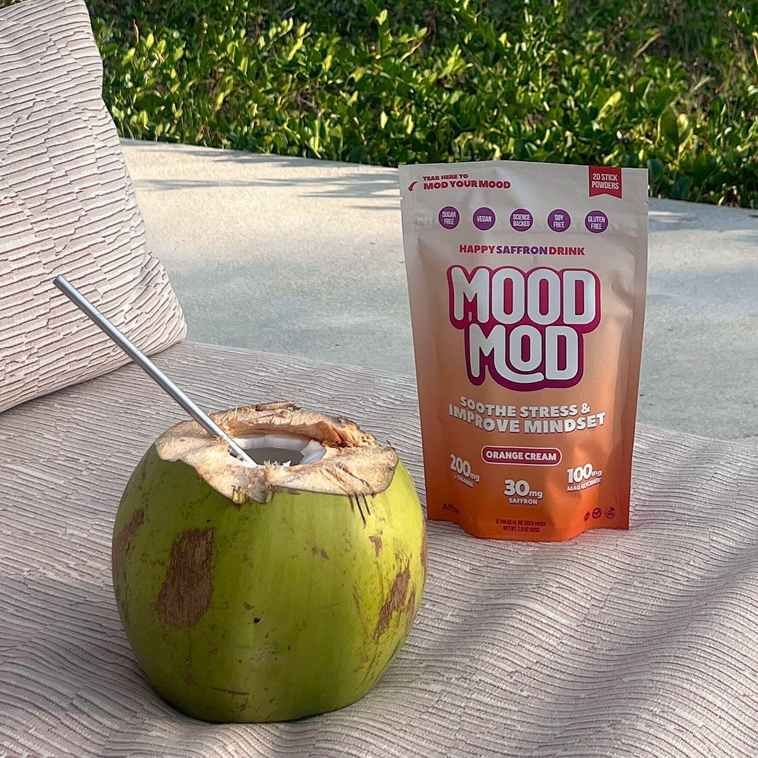 Mood Mod Orange Cream saffron drink pouch next to a fresh coconut with a metal straw on an outdoor lounge chair