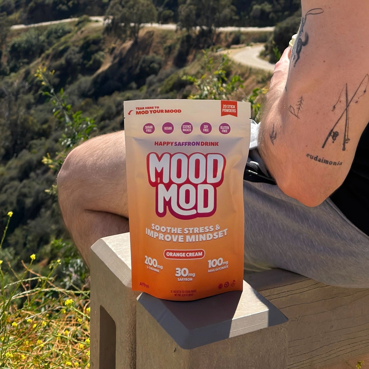 Mood Mod Orange Cream saffron drink pouch resting on a wooden railing outdoors with a hillside trail and wildflowers in the background