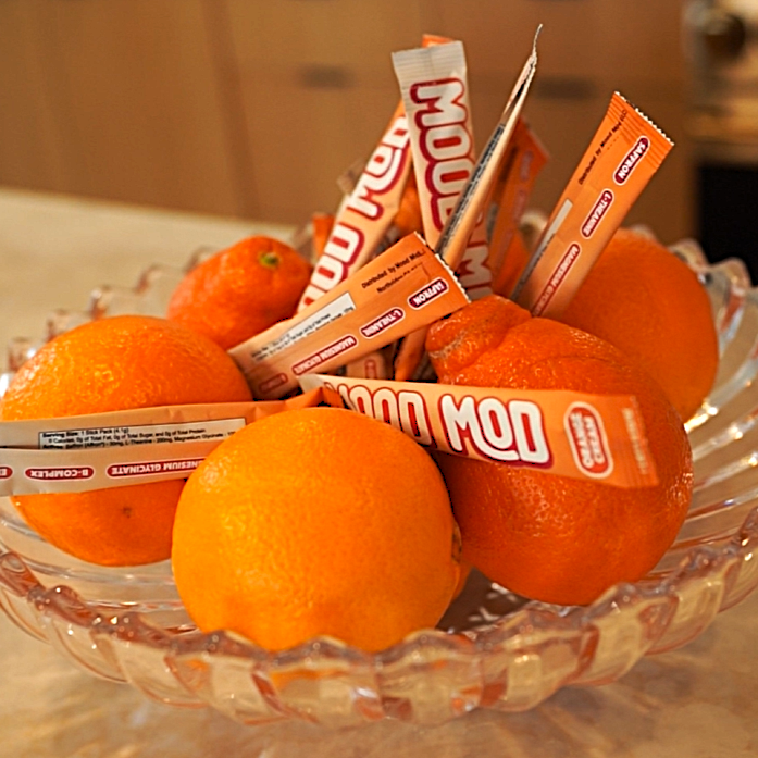 Mood Mod orange-flavored stick packs displayed in a glass bowl alongside fresh oranges on a kitchen countertop.