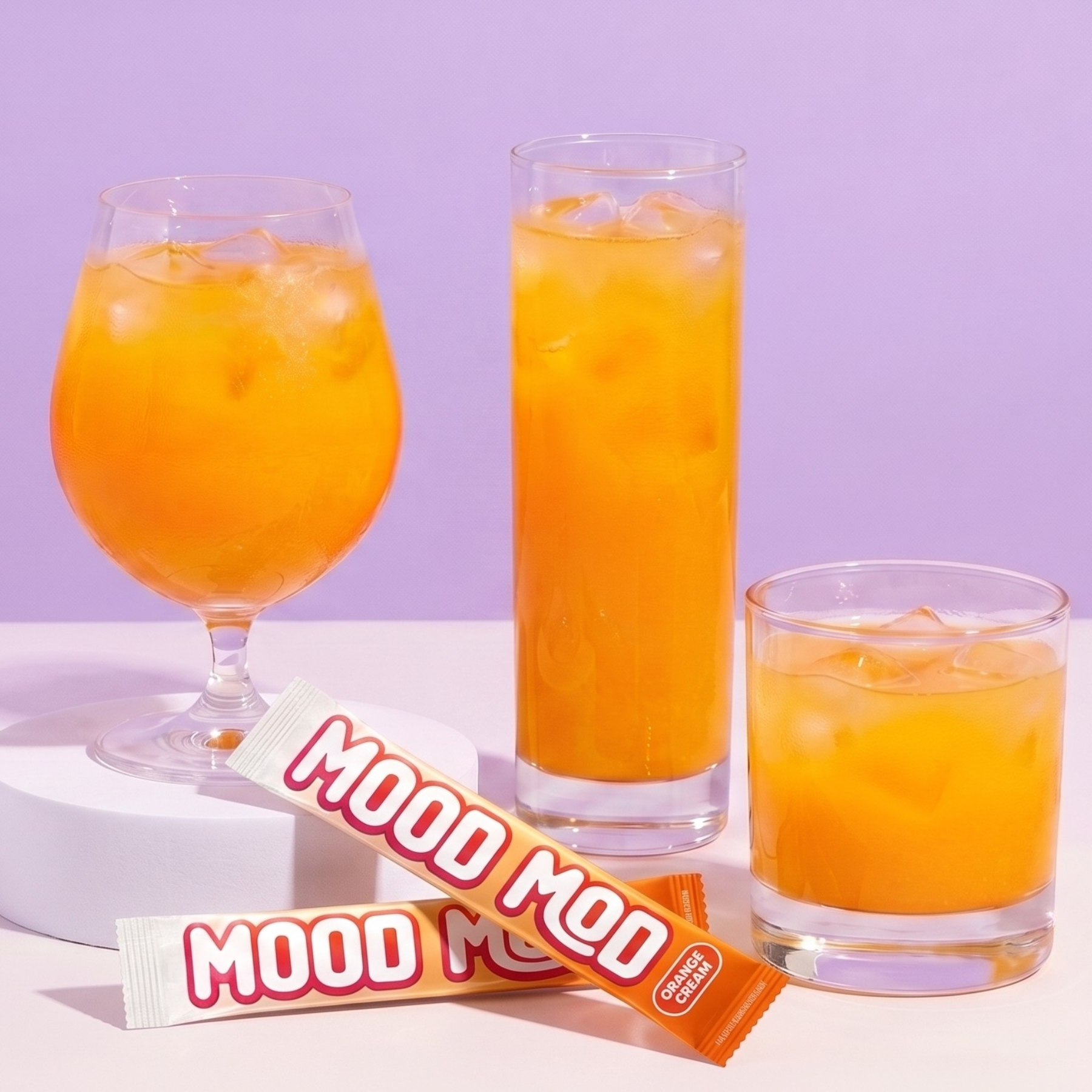 Three glasses of Mood Mod Orange Cream drink over ice next to two single-serve stick packets on a lavender background