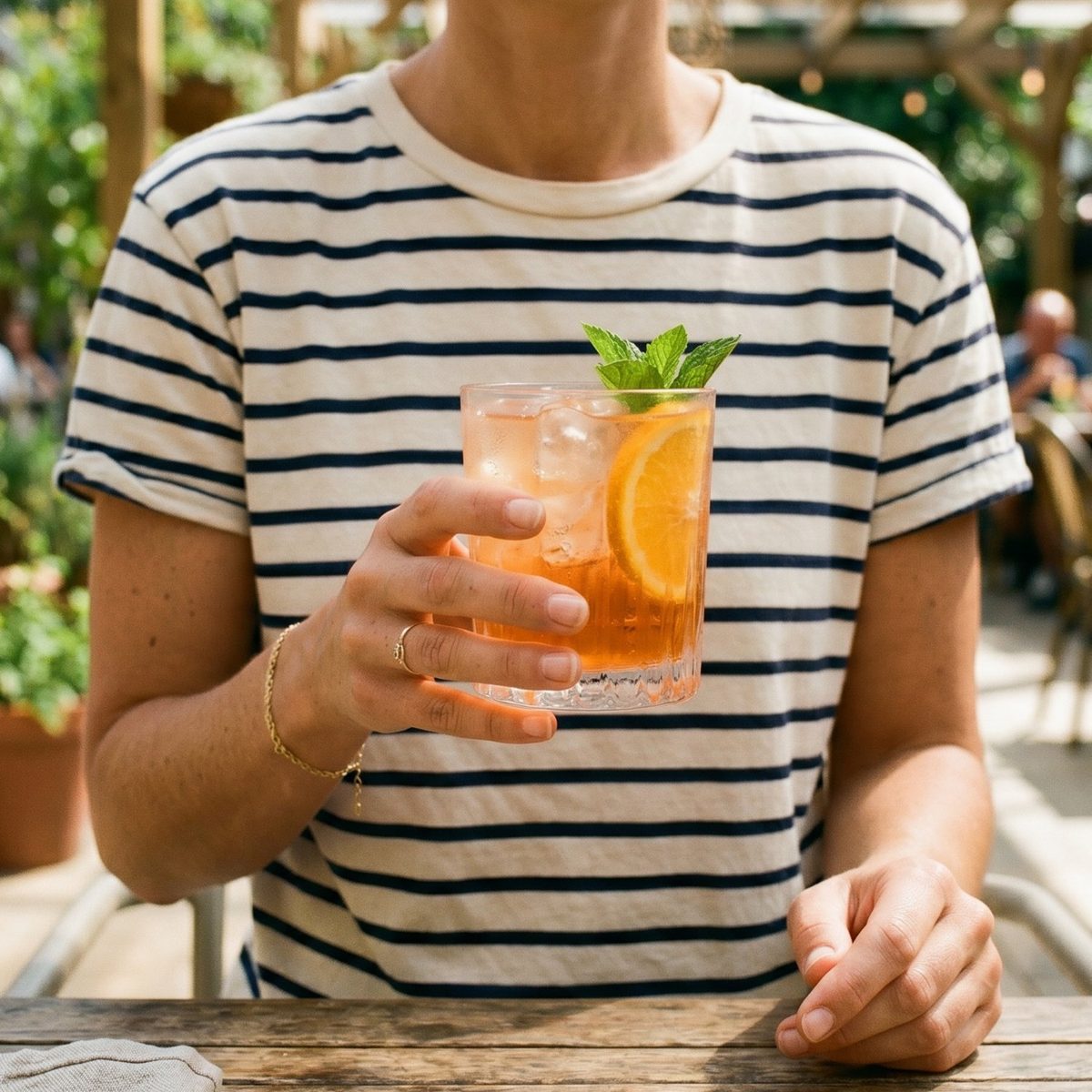 Person in a striped t-shirt holding a Mood Mod orange drink with mint and an orange slice at an outdoor café.