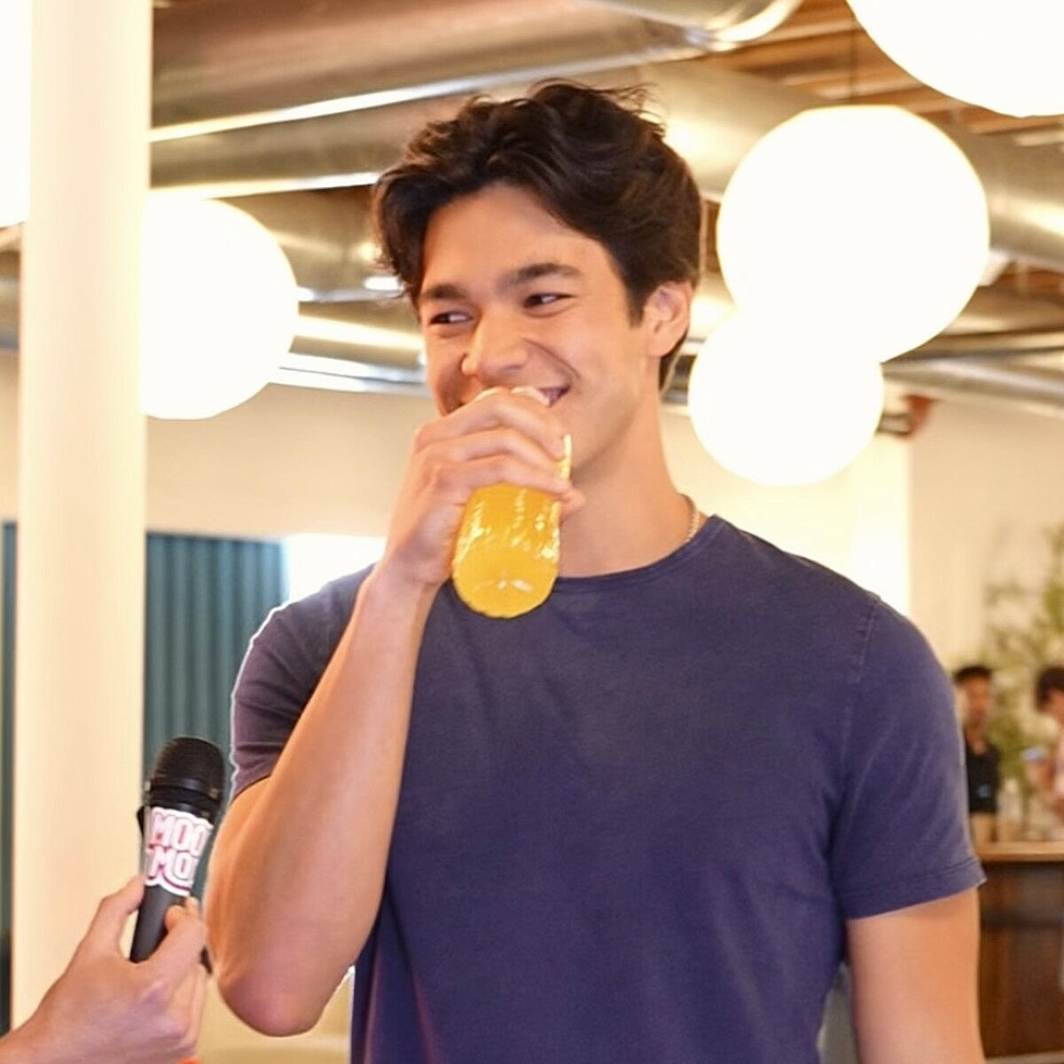 Young man in a navy t-shirt smiling as he drinks an orange Mood Mod supplement stick in a bright modern office.