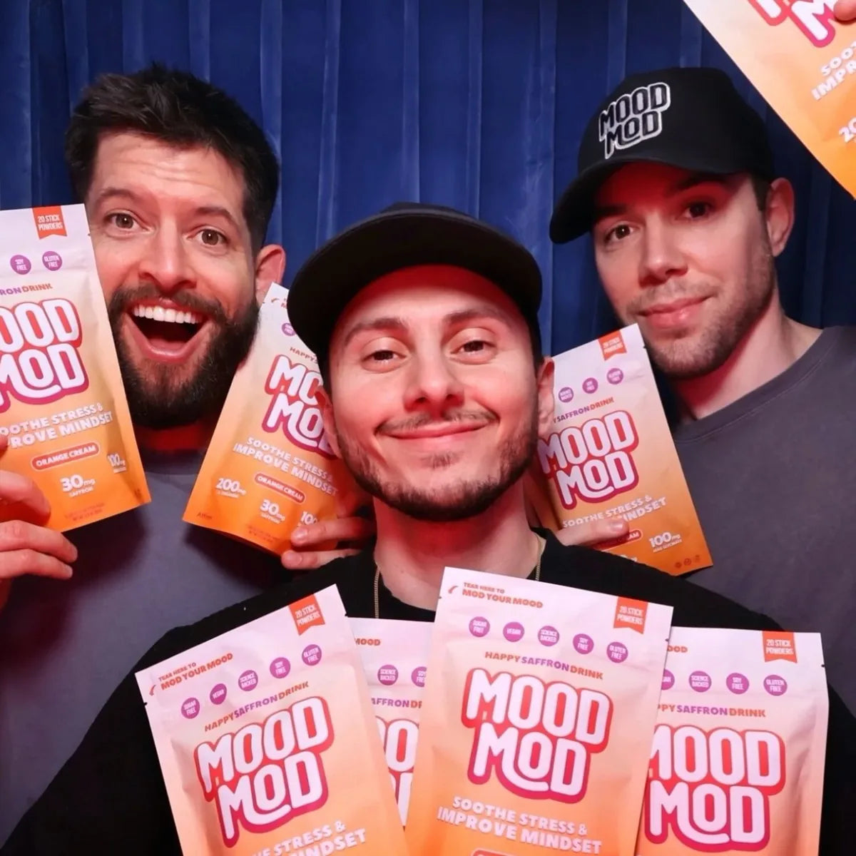 Mood Mod Happy Saffron Drink team holding orange and pink stick powder packets featuring 30mg saffron extract, 200mg L-theanine, and 100mg magnesium glycinate for stress relief and improved mindset