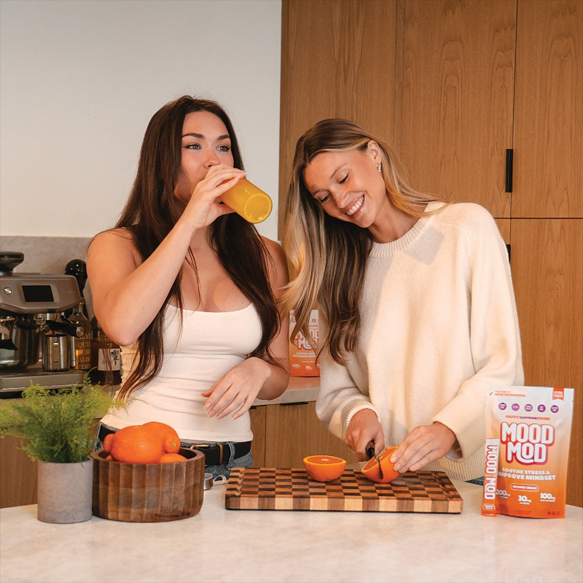 Smiling woman cutting a fresh orange next to a Mood Mod saffron drink mix pouch, while a friend drinks an orange beverage in a bright modern kitchen.