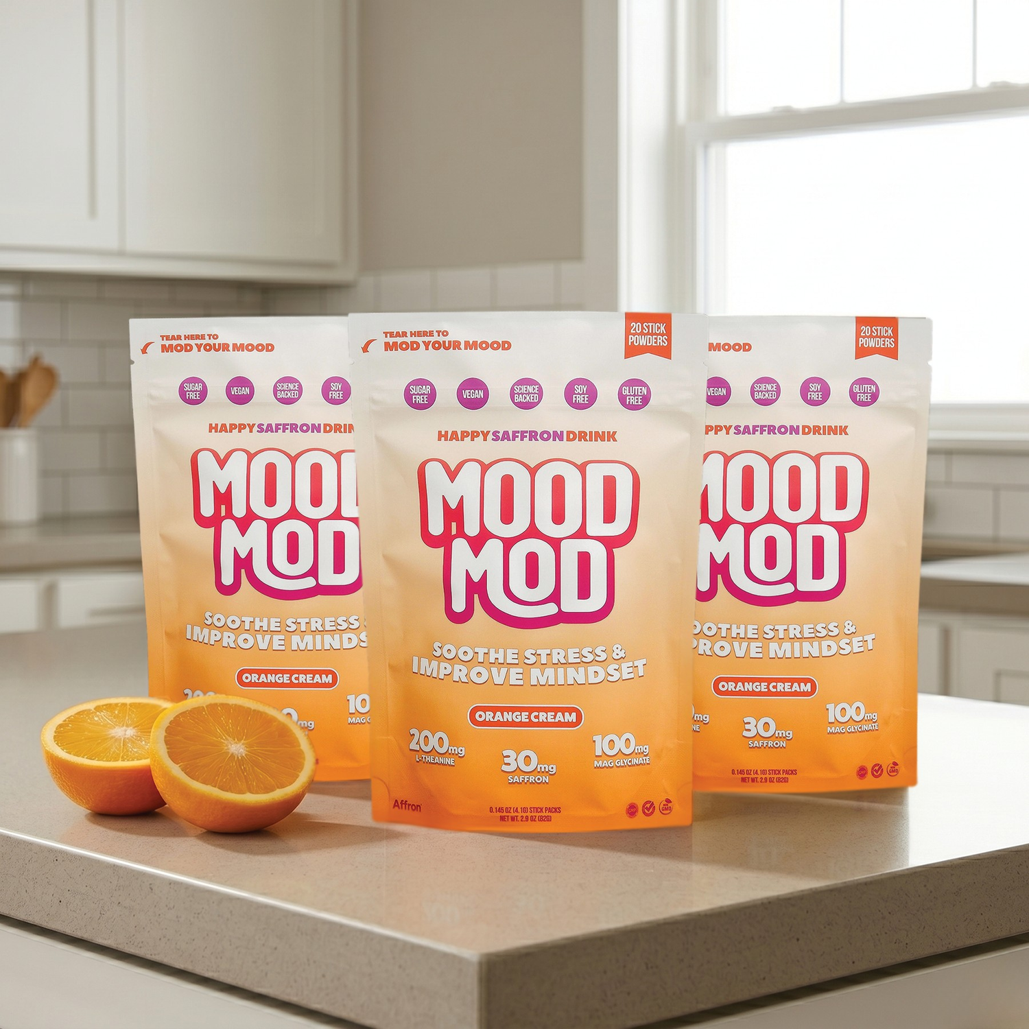Three pouches of "MOOD MOD" Happy Saffron Drink in Orange Cream flavor are displayed on a light gray kitchen countertop next to two halved oranges. The packaging features bold pink and white text that reads "Soothe Stress & Improve Mindset" and lists key ingredients: 200mg L-theanine, 30mg Saffron, and 100mg Magnesium Glycinate. The background shows a bright, modern kitchen with white cabinets and a window.