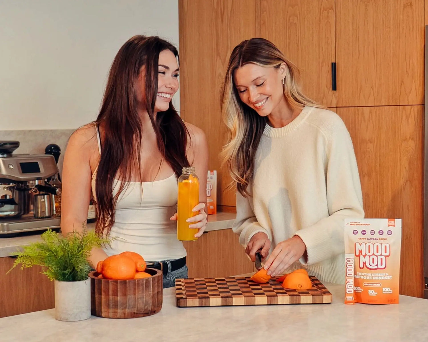Two women smiling in a modern kitchen, one holding a bottle of orange juice while the other slices fresh oranges on a wooden cutting board next to a bag of Mood Mod Happy Saffron Drink in Orange Cream flavor.